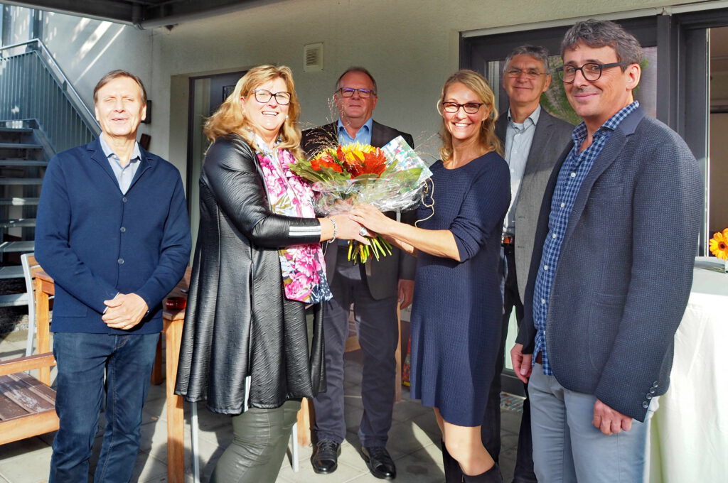 Eine Gruppe von Menschen lächelt in die Kamera. Ein großer Blumenstrauß wird überreicht.