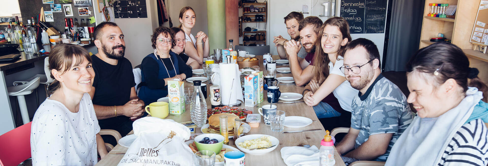 Zehn junge Bewohner einer inklusiven WG sitzen um den gedeckten Frühstückstisch herum. Alle lächeln freundlich in die Kamera.