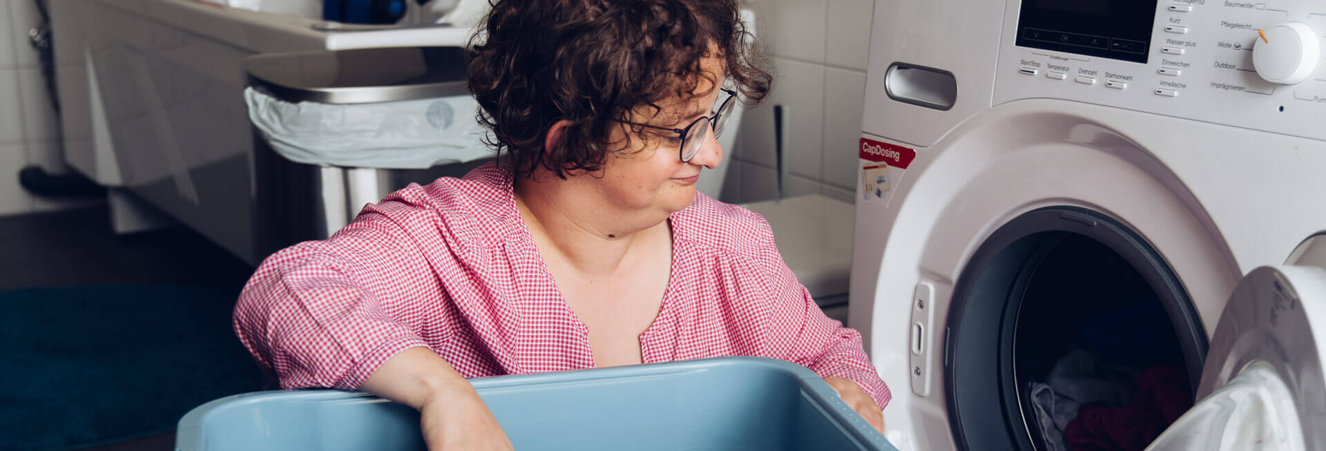 Ein Mädchen mit Behinderung hockt mit einem Wäschekorb vor der geöffneten Waschmaschine.