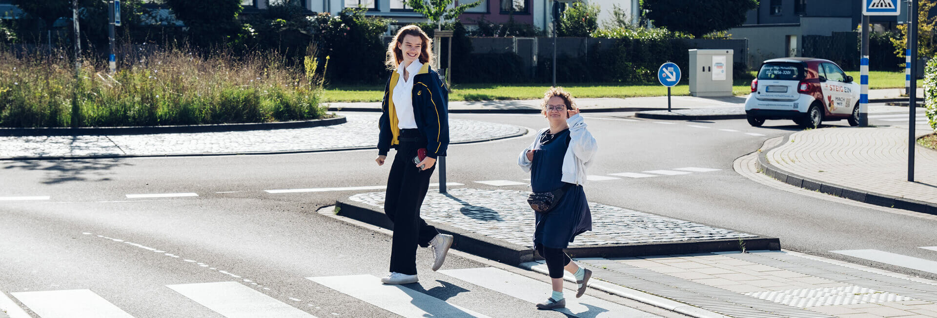 Zwei Mädchen, mit und ohne Behinderung, überqueren gemeinsam die Straße, wobei sie lachen und in die Kamera winken.