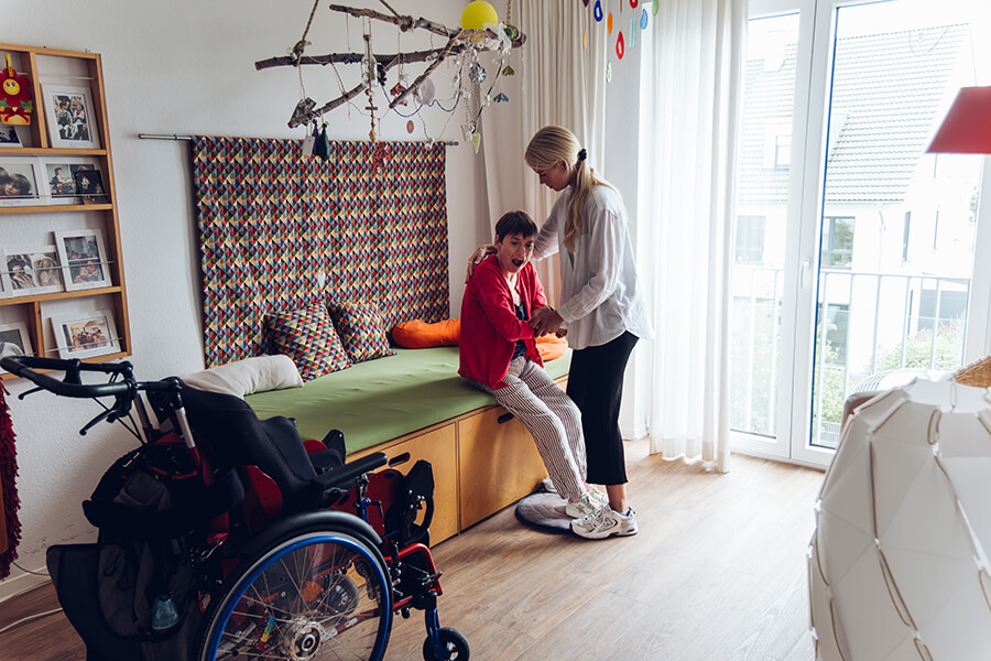 Ein Mädchen ohne Behinderung unterstützt ein Mädchen mit Behinderung dabei, vom Bett aufzustehen und sich in einen Rollstuhl zu setzen.