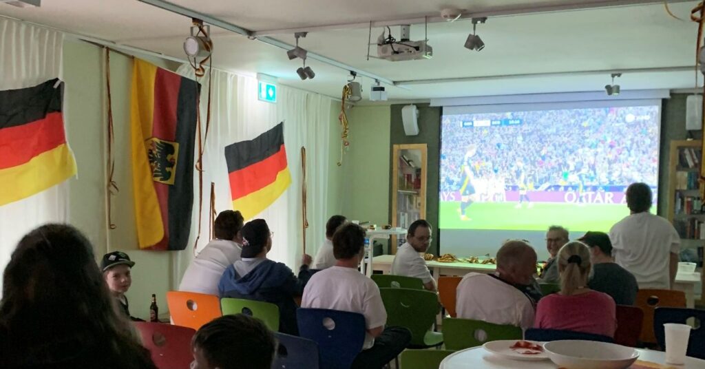 Einige Menschen mit und ohne Behinderung sitzen in einem abgedunkelten Raum vor einer Leinwand, auf der ein Fußballspiel läuft. An der Wand hängen Deutschlandflaggen.